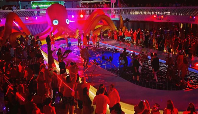 A wide-shot of a pool party with many people dancing, most dressed in red. A large inflatable octopus sits behind the pool