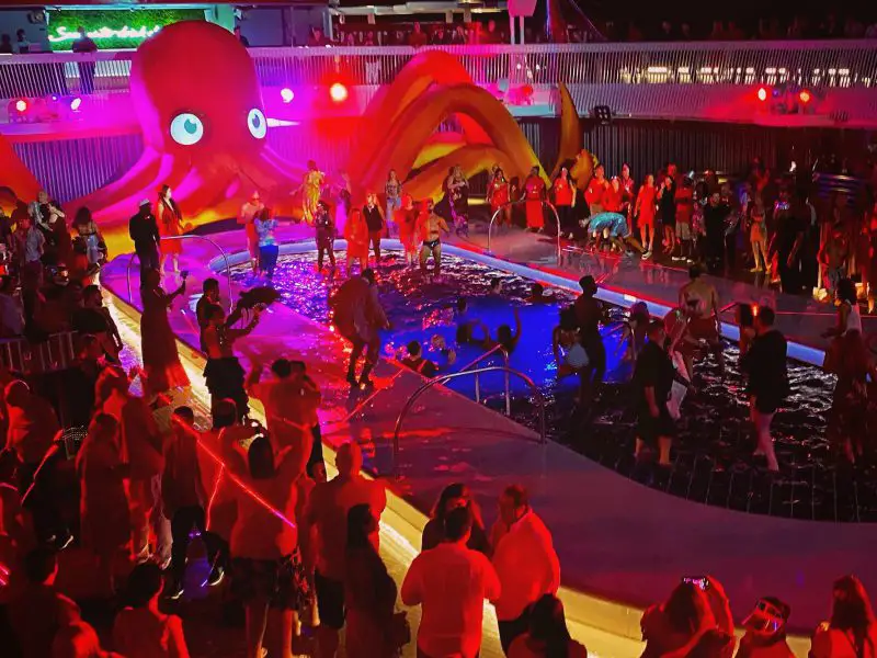 A wide-shot of a pool party with many people dancing, most dressed in red. A large inflatable octopus sits behind the pool