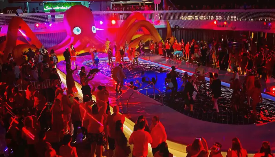 A wide-shot of a pool party with many people dancing, most dressed in red. A large inflatable octopus sits behind the pool