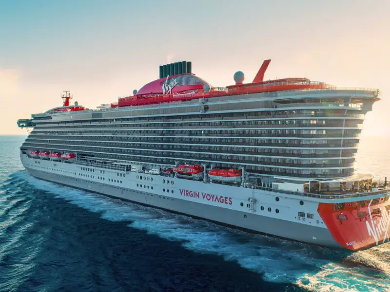 A large grey cruise ship cutting through the waves with red lifeboats and funnels. The ship is set against a setting sun backdrop.