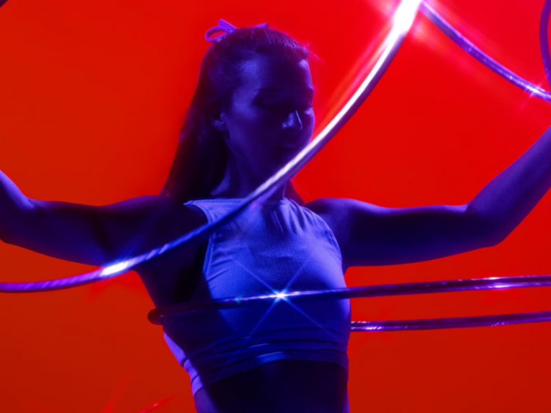 A woman spinning 5 hula hoops lit in blue, with a red backdrop