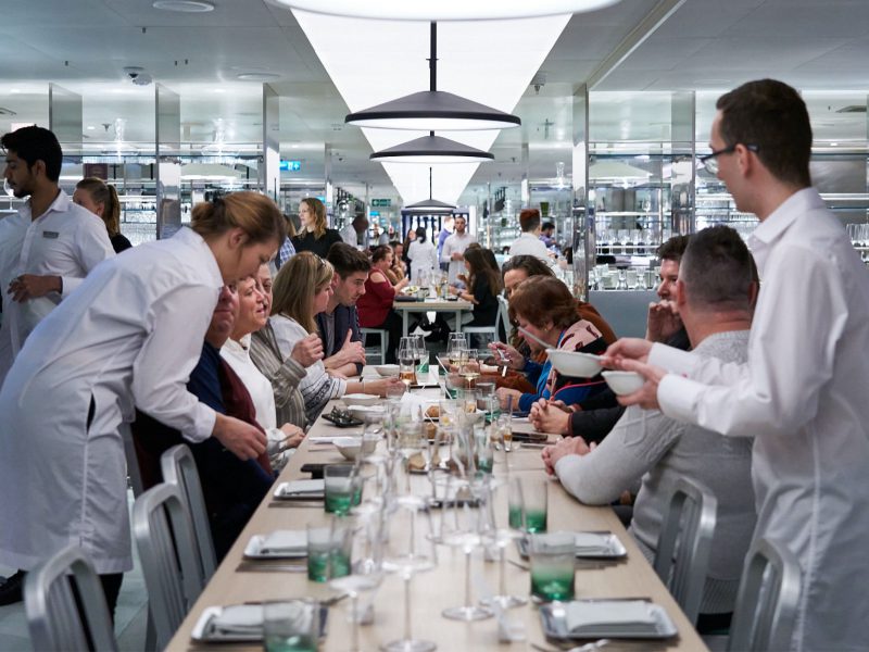 A number of people eating dinner in a restaurant designed to look like a science lab, diners are gathered around a long table with waiters in lab coats attending to them