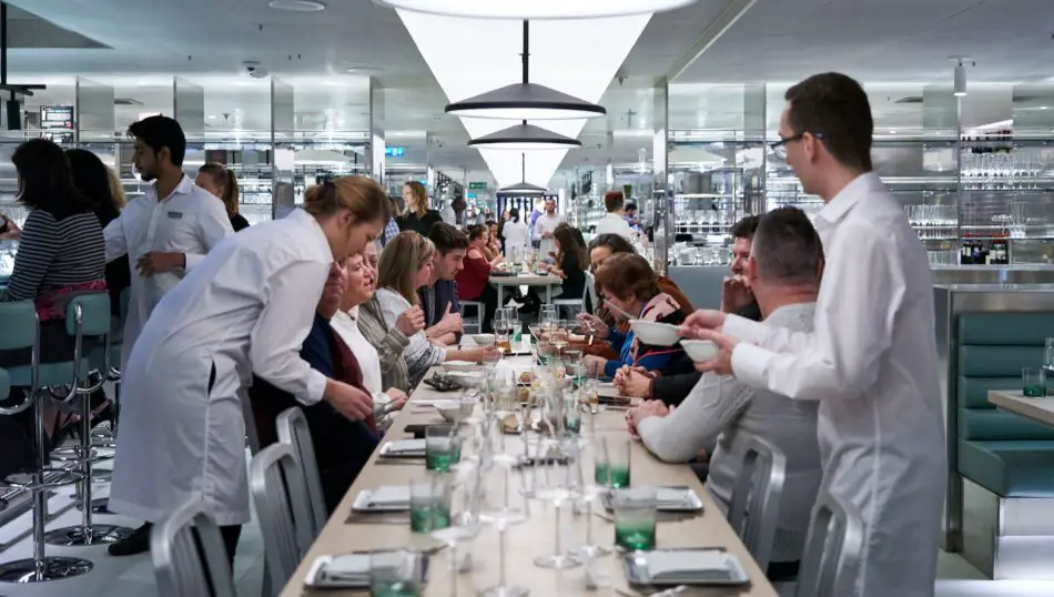 A number of people eating dinner in a restaurant designed to look like a science lab, diners are gathered around a long table with waiters in lab coats attending to them