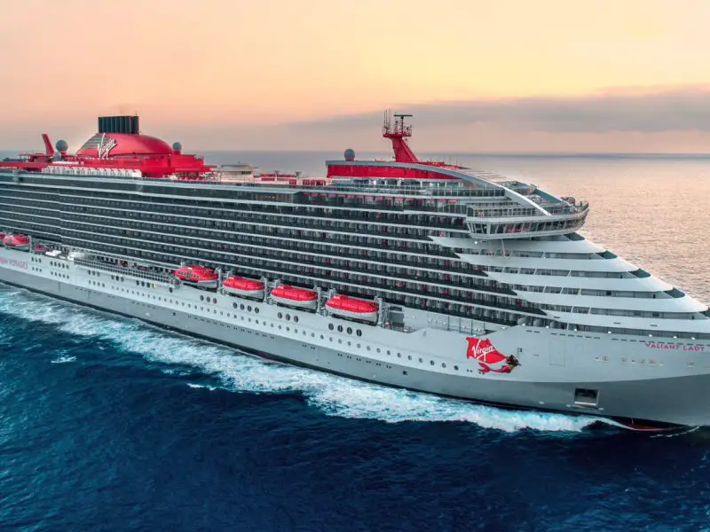 A large grey cruise ship cutting through the waves with red lifeboats and funnels. There is a glamorous mermaid painted on the side.