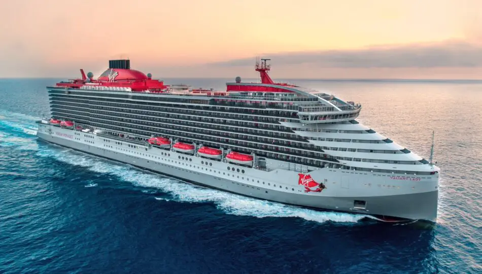 A large grey cruise ship cutting through the waves with red lifeboats and funnels. There is a glamorous mermaid painted on the side.