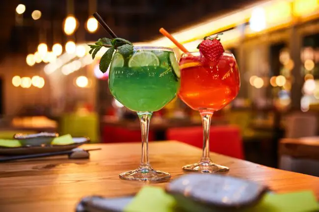 Two colourful cocktail drinks against a brightly lit bar background
