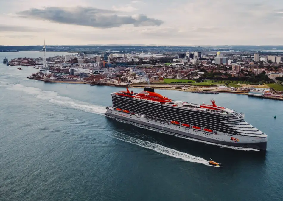 A Virgin Voyages cruise ship sails past the coastline of Portsmouth with an orange tug boat sailing alongside