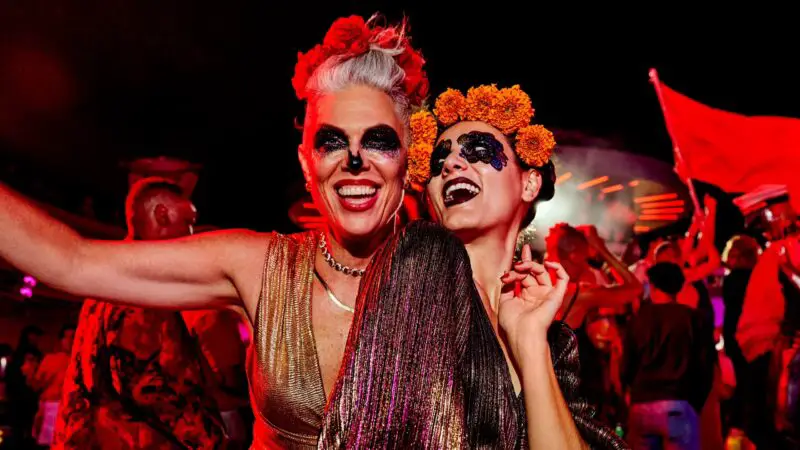 Two people dance with skeleton face paint wearing long sparkling dresses in an outdoor settings under bright lights at night.