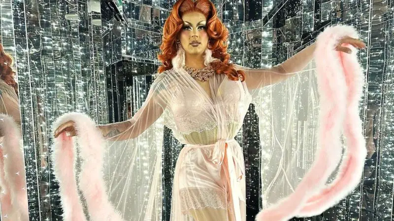 a drag queen in a long white dress with large sleeves posing in a glittering hallway