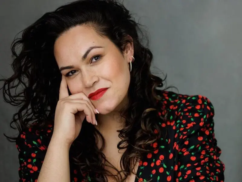 A woman with long dark curly hair wearing a red pattern shirt against a grey background