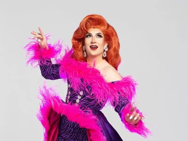 A drag queen wearing a pink and purple feather-lined dress against a white background