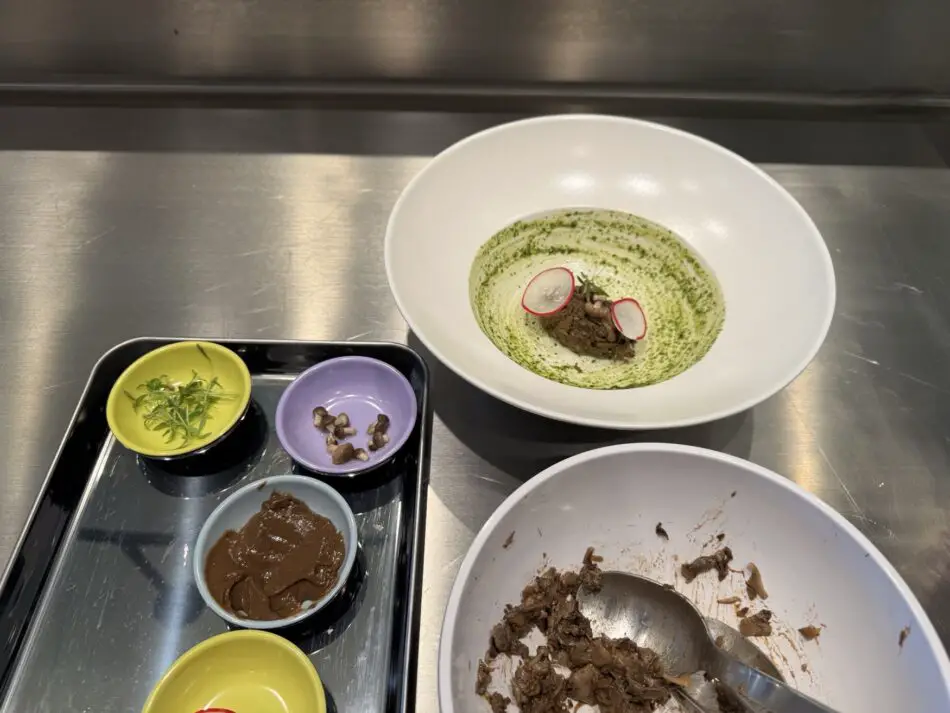 Large white bowl with green herb sauce and meat, radish slices, beside a metal tray of colorful condiment bowls
