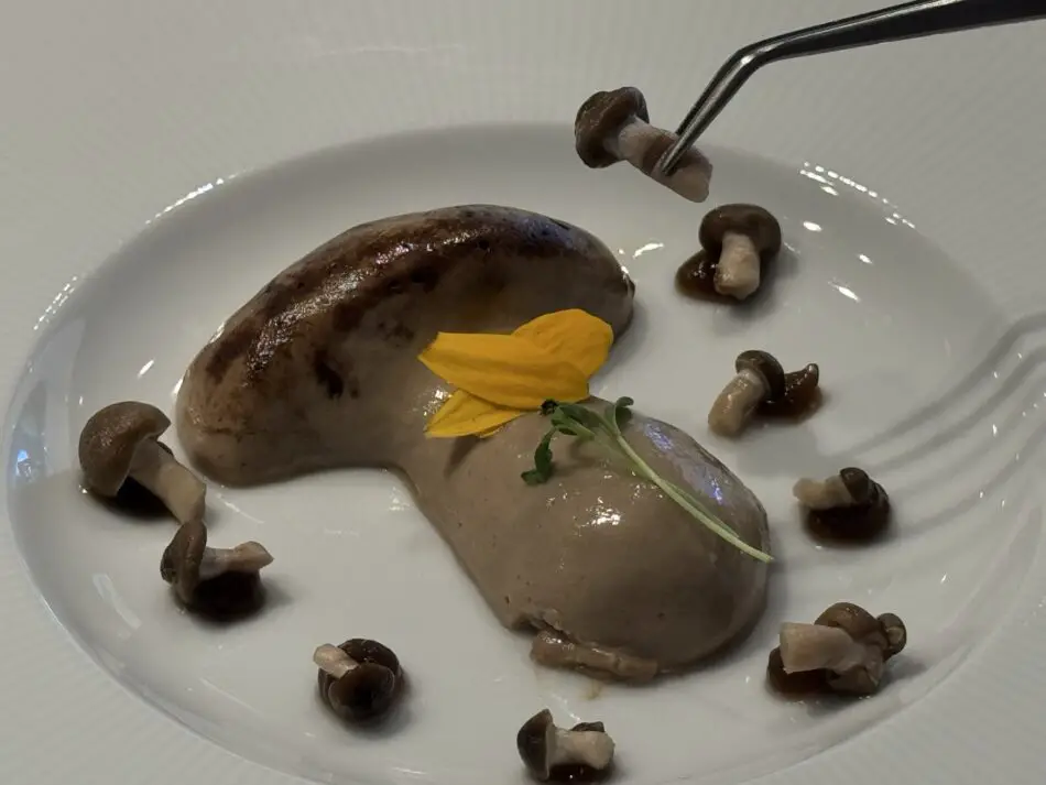 Two brown mousse quenelles with mushroom garnish and a yellow edible flower on a white plate