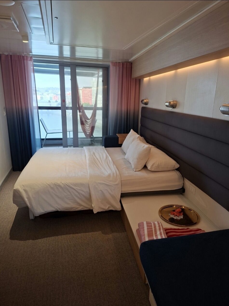 Virgin Voyages cruise cabin with a large bed and balcony with hammock and city view