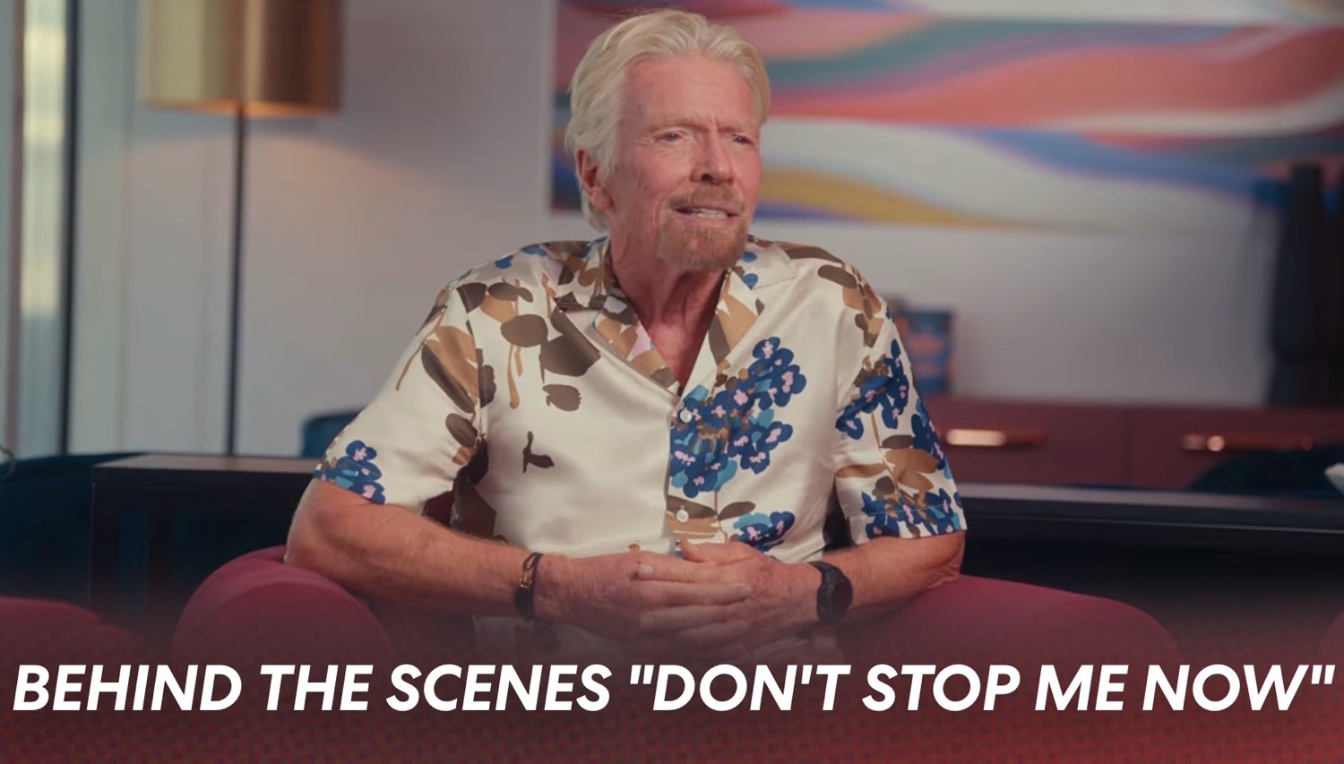 Man with white hair in a floral shirt sits on a red couch in a modern room