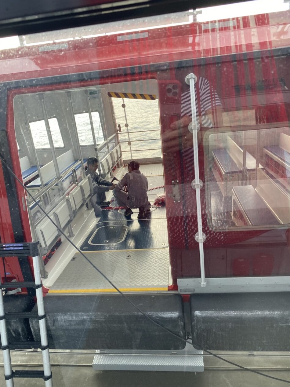 Two people sitting on the deck of a red Virgin Voyages cruise ship with water in the background