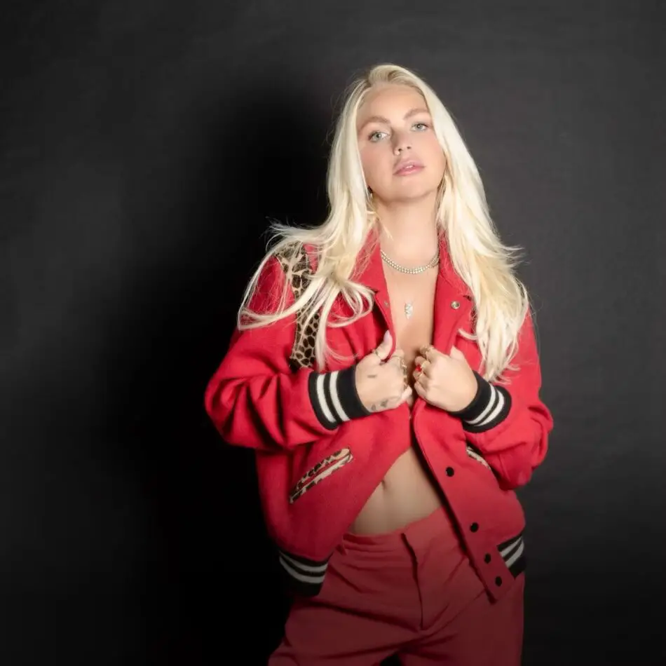 Blonde woman wearing a red varsity jacket with leopard patches posing against a dark backdrop
