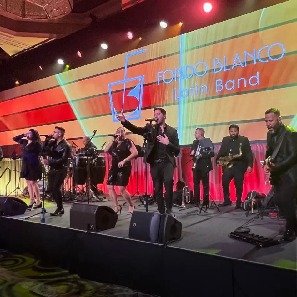 Latin band performing on stage with colorful backdrop reading Fondo Blanco Latin Band