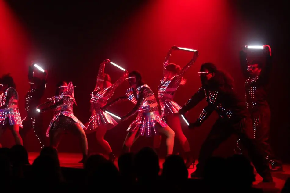 Dancers in LED-lit outfits perform on a red-lit stage, holding glowing light bars