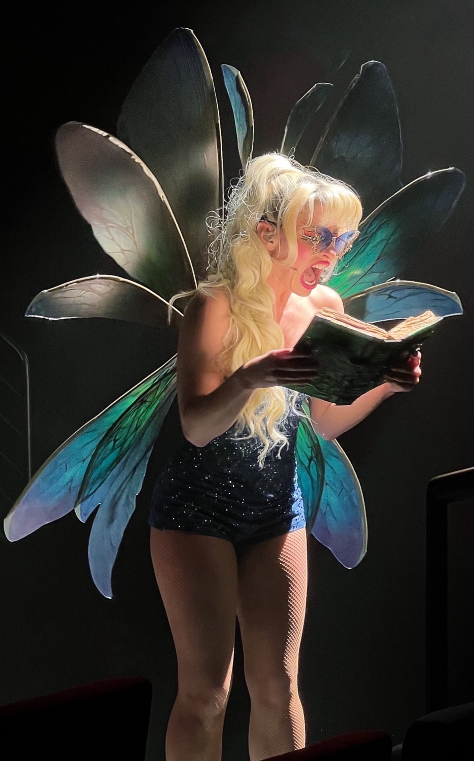 Performer in a sparkling blue outfit with large butterfly wings reads a book on stage
