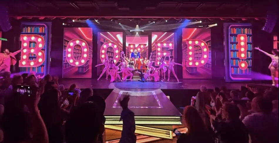 Stage show in a cruise ship theater with performers in pink outfits and large illuminated BOOKED letters, audience watching