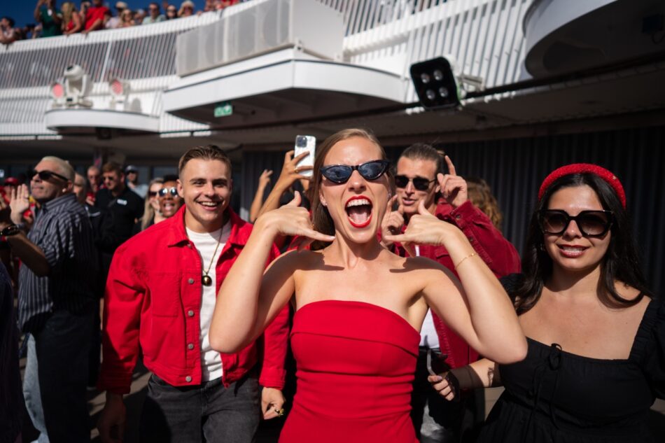 Crowd on a Virgin Voyages cruise ship deck celebrating, central woman in red dress shouting and posing