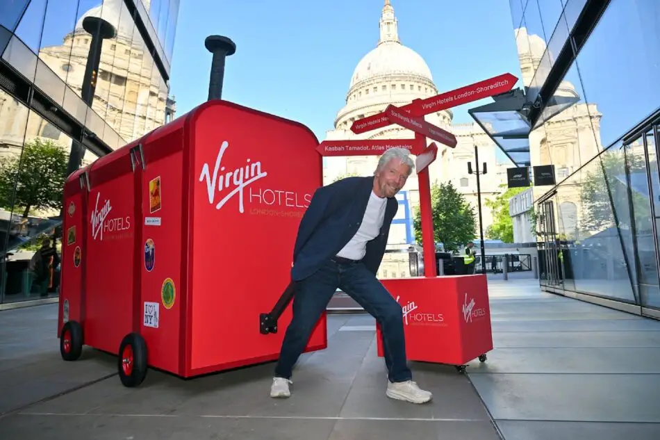 Richard Branson posing next to a large red Virgin Hotels trailer in London