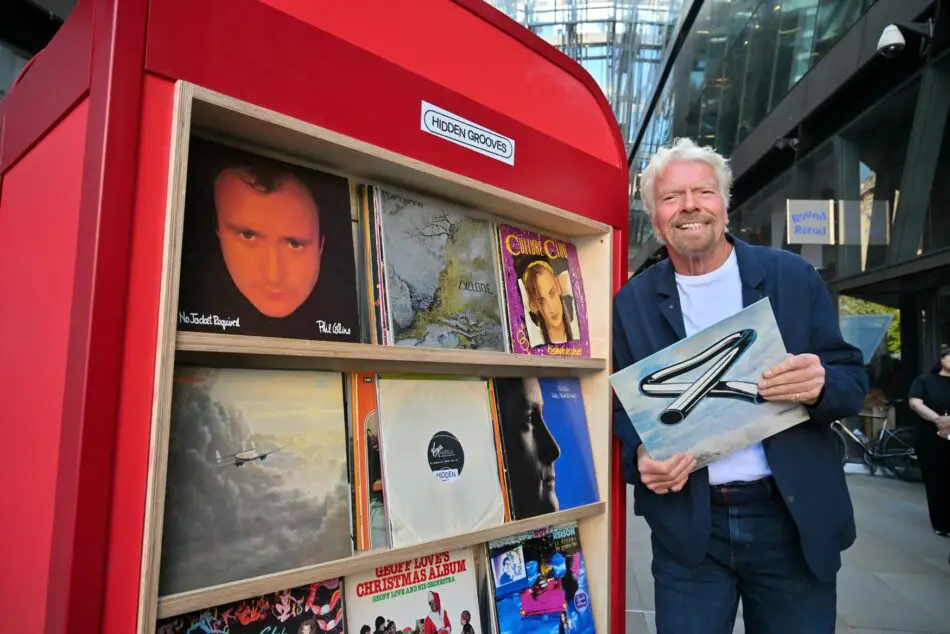 Richard Branson stands beside a red outdoor vinyl record display holding a vinyl album cover
