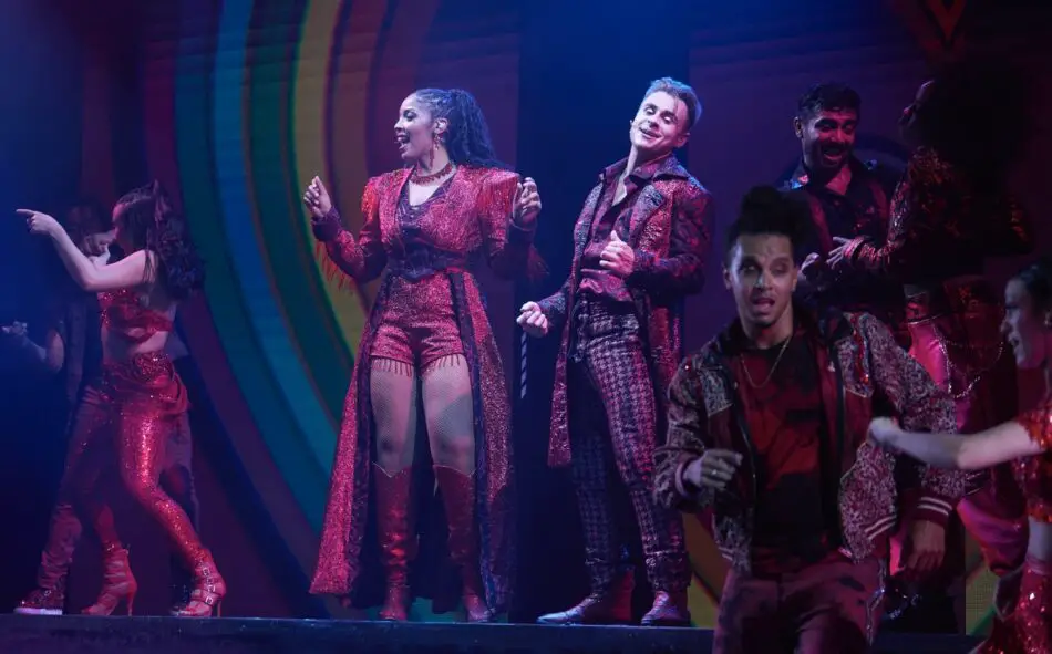 Dancers in red sequined outfits perform on a colorful stage with rainbow arches