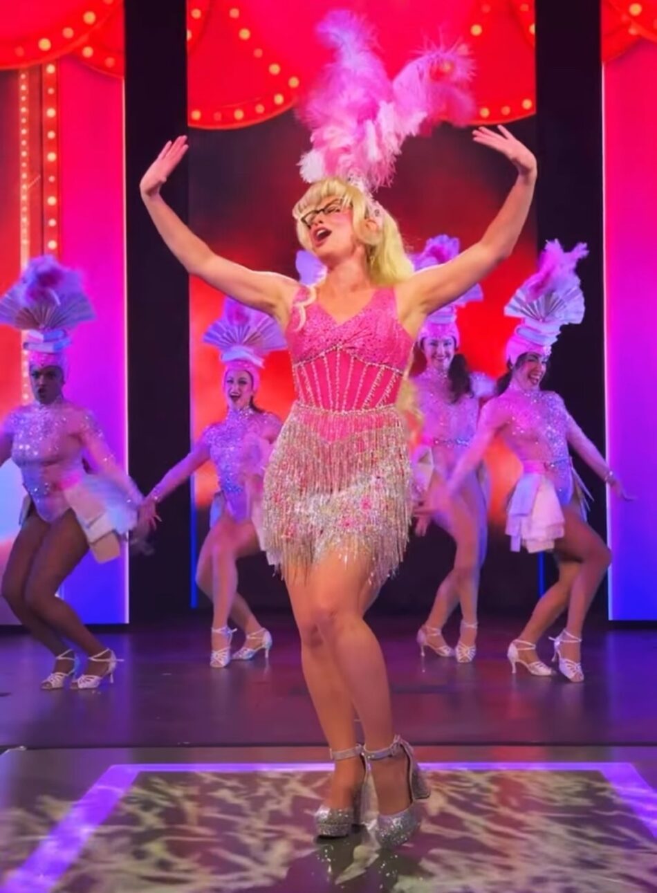 Blonde performer in a pink fringe dress with feathered headdress performs on stage with backup dancers