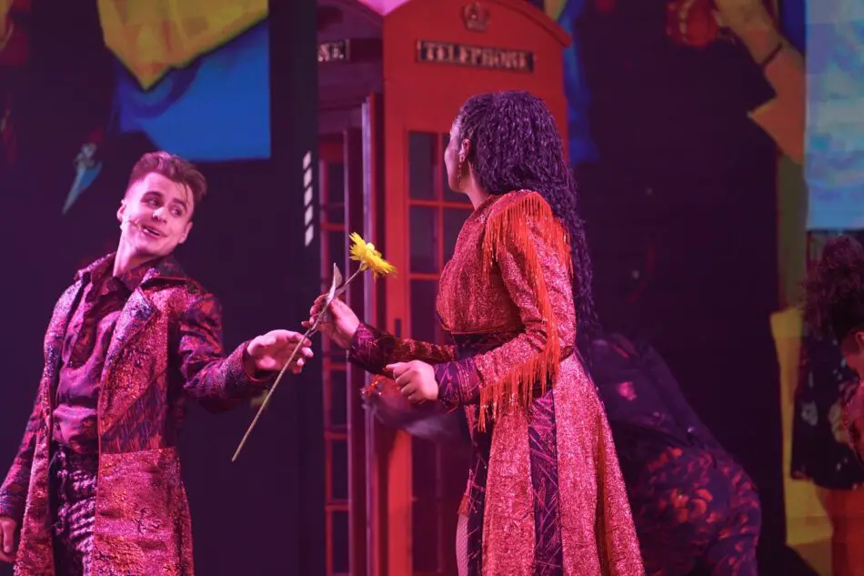 Two performers in glittering red costumes on stage, handing a sunflower near a red telephone booth
