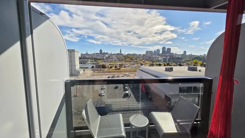 Balcony cabin with two white chairs and a small table overlooking a city skyline on a Virgin Voyages cruise ship