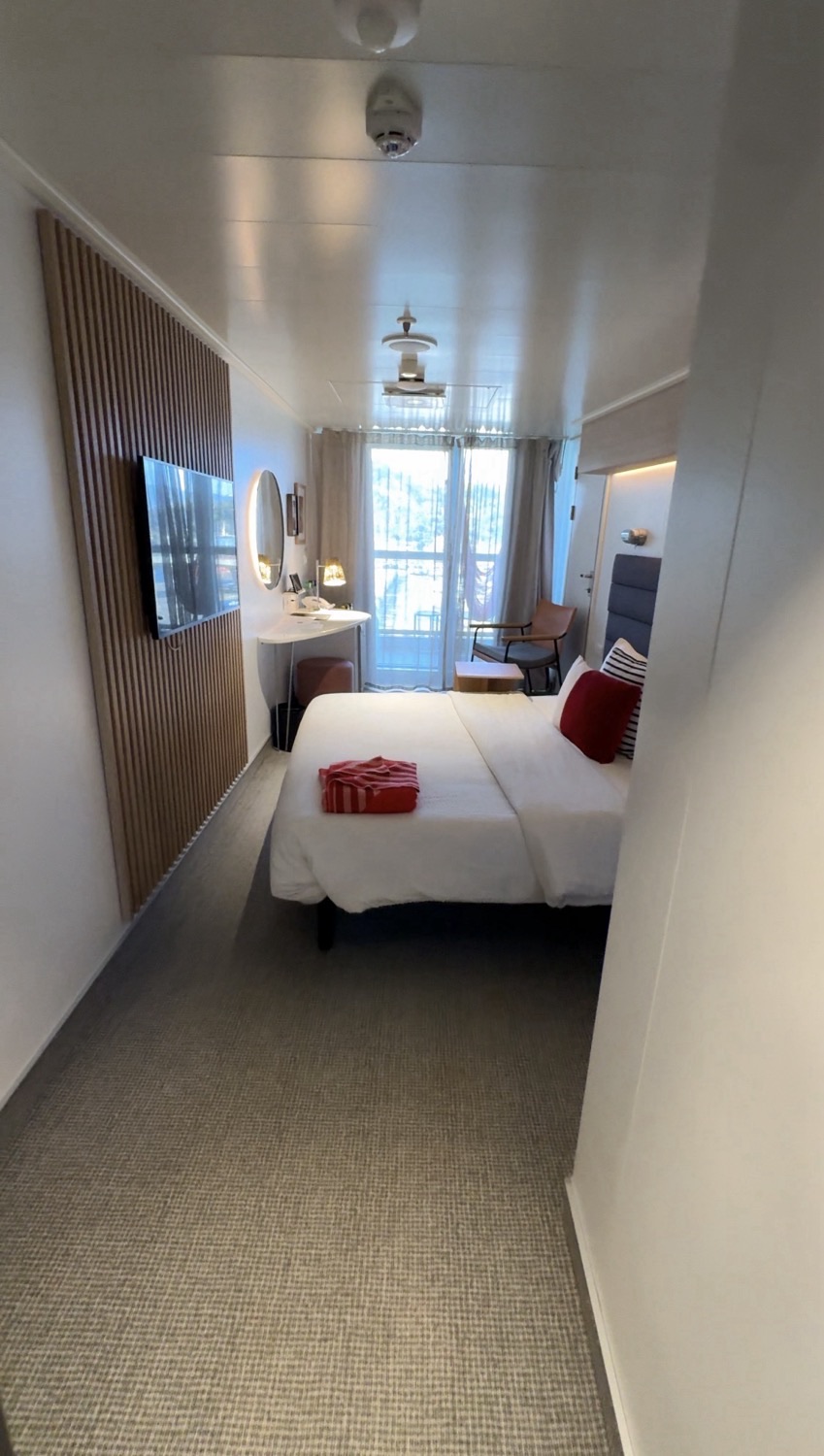 Virgin Voyages cruise ship cabin with bed, wall TV, desk, and balcony