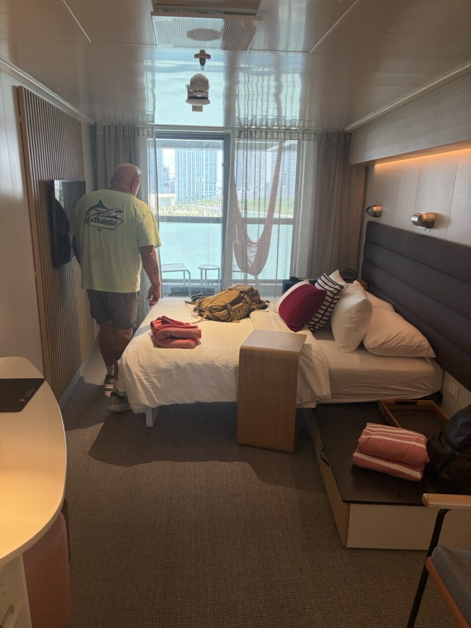 Man in a light shirt stands beside a bed in a Virgin Voyages cruise ship cabin, balcony view