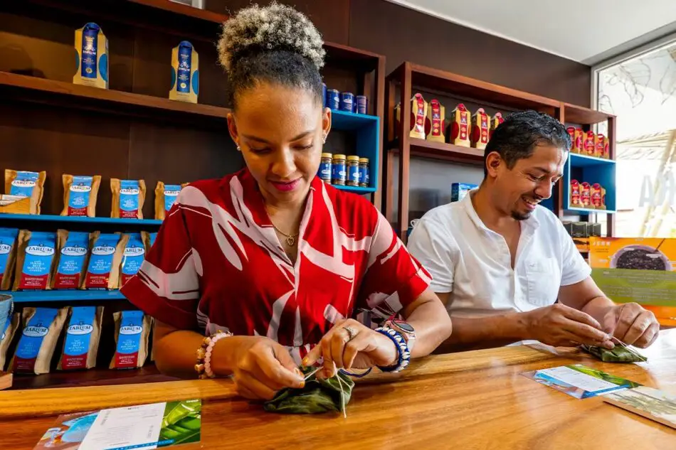 Two people wrap green leaves at a wooden counter in a shop
