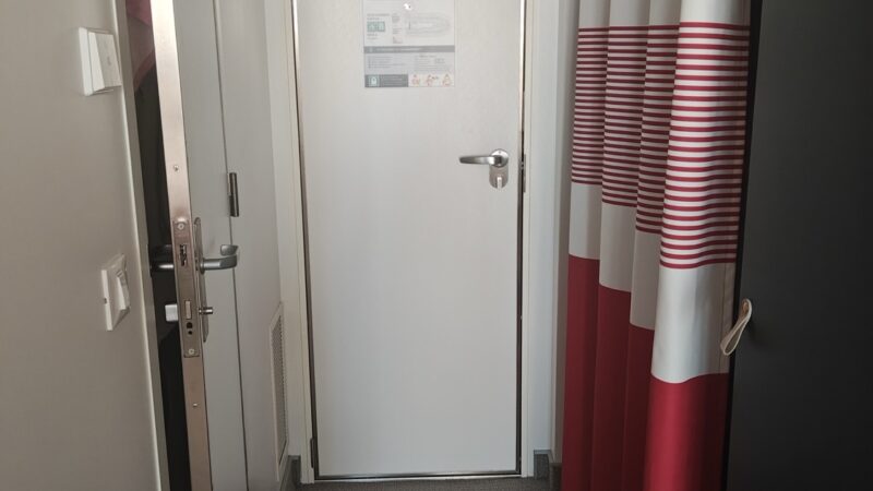 Narrow cabin corridor with a white door and red striped curtain on a Virgin Voyages cruise ship