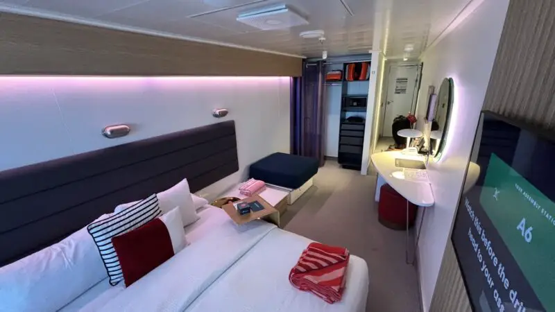 Interior of a Virgin Voyages cruise ship cabin with a large bed and built-in desk