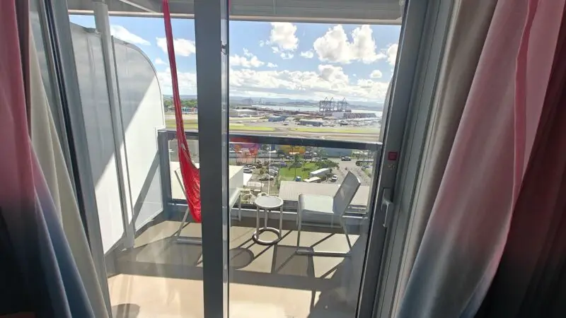 Balcony of a Virgin Voyages cruise ship cabin with red hammock and outdoor seating overlooking the port