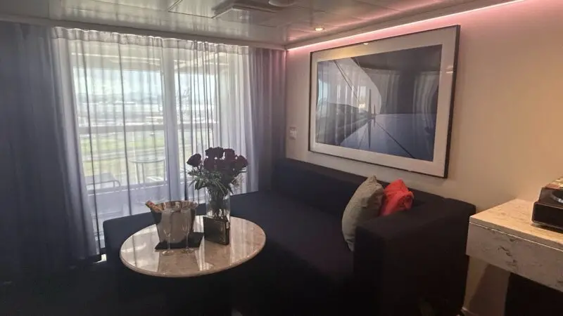 Living area of a Virgin Voyages cruise ship cabin with sofa, round table, flowers, and balcony view