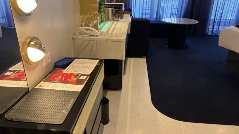 Cruise cabin with marble minibar, mirror, phone, and blue drapes on a Virgin Voyages ship