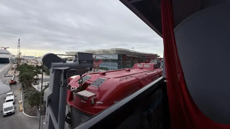 View from a Virgin Voyages cabin balcony of a red lifeboat on deck with harbor in background