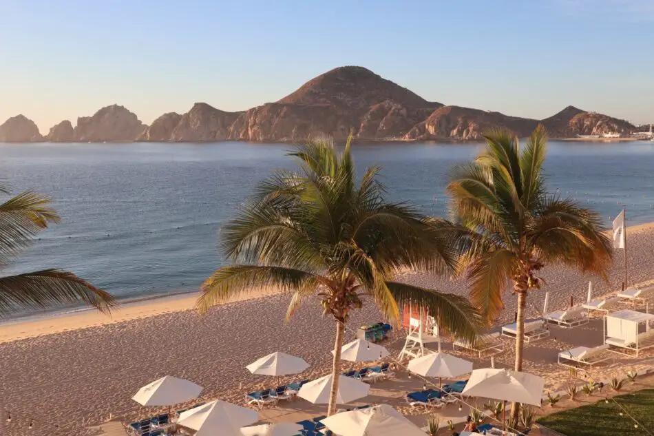 Sunny beach with palm trees, white umbrellas, and a distant rocky island