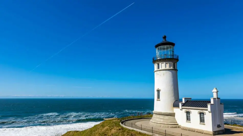 White lighthouse on a grassy cliff overlooking the ocean with a clear blue sky and visible contrail.