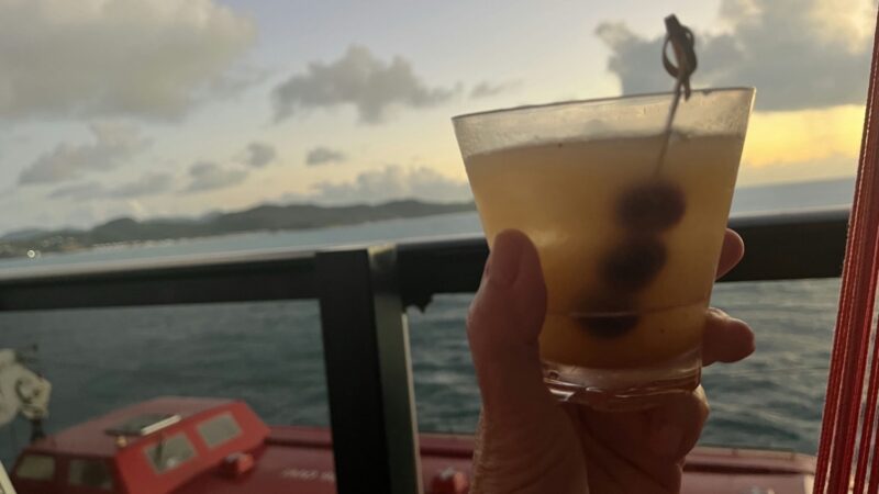 A hand holds a cocktail with olives on a Virgin Voyages cruise ship at sunset