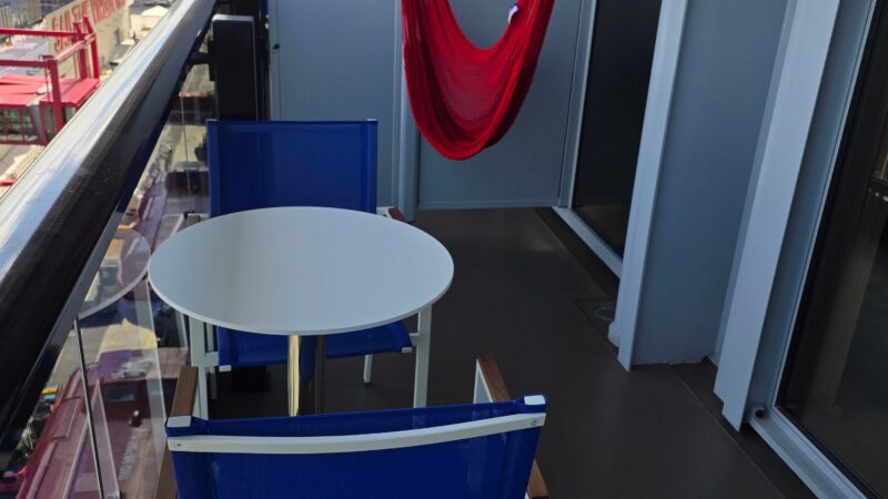 Virgin Voyages cruise ship cabin balcony with a red hammock, blue chairs, a round table, and city view in the background.
