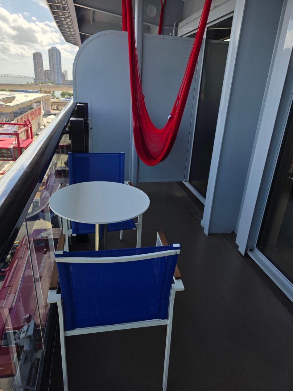 Virgin Voyages cruise ship cabin balcony with a red hammock, blue chairs, a round table, and city view in the background.