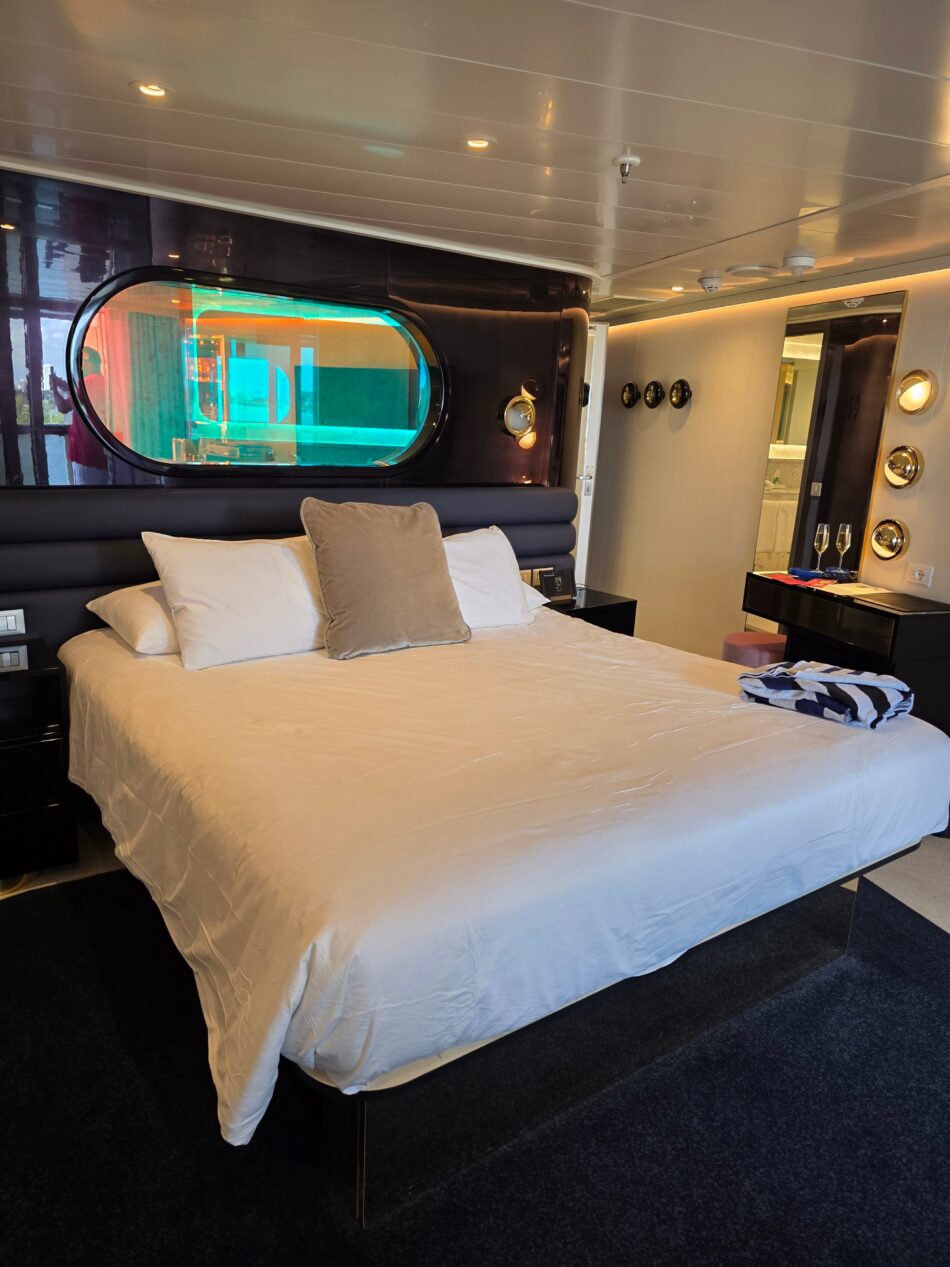 Cruise ship cabin with a large bed, oval window, modern lighting, and reflective surfaces.