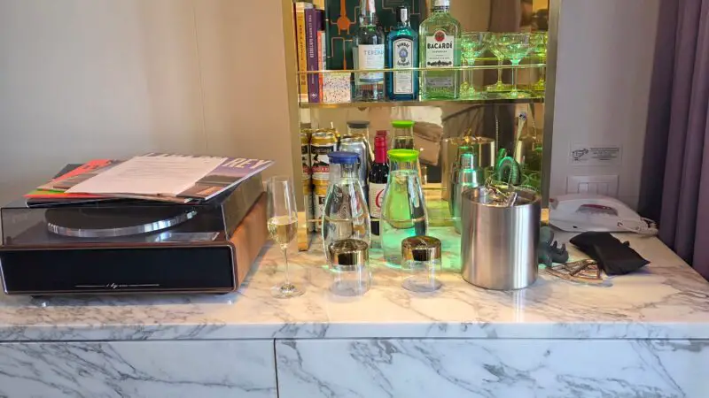 Virgin Voyages cruise ship cabin bar area featuring a marble countertop with a record player, glasses, and bottles; backlit shelves hold liquor and more glassware.