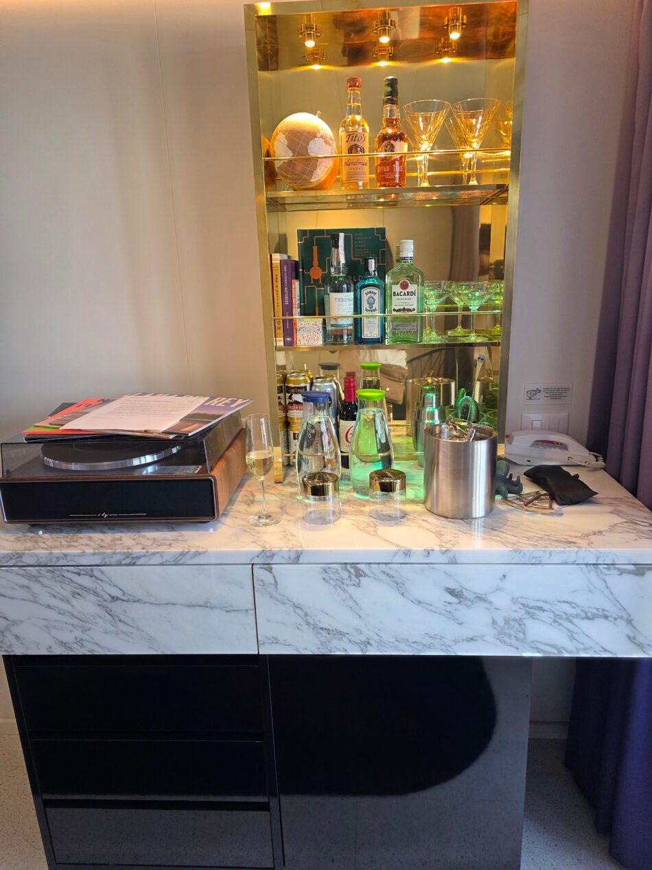 Virgin Voyages cruise ship cabin bar area featuring a marble countertop with a record player, glasses, and bottles; backlit shelves hold liquor and more glassware.