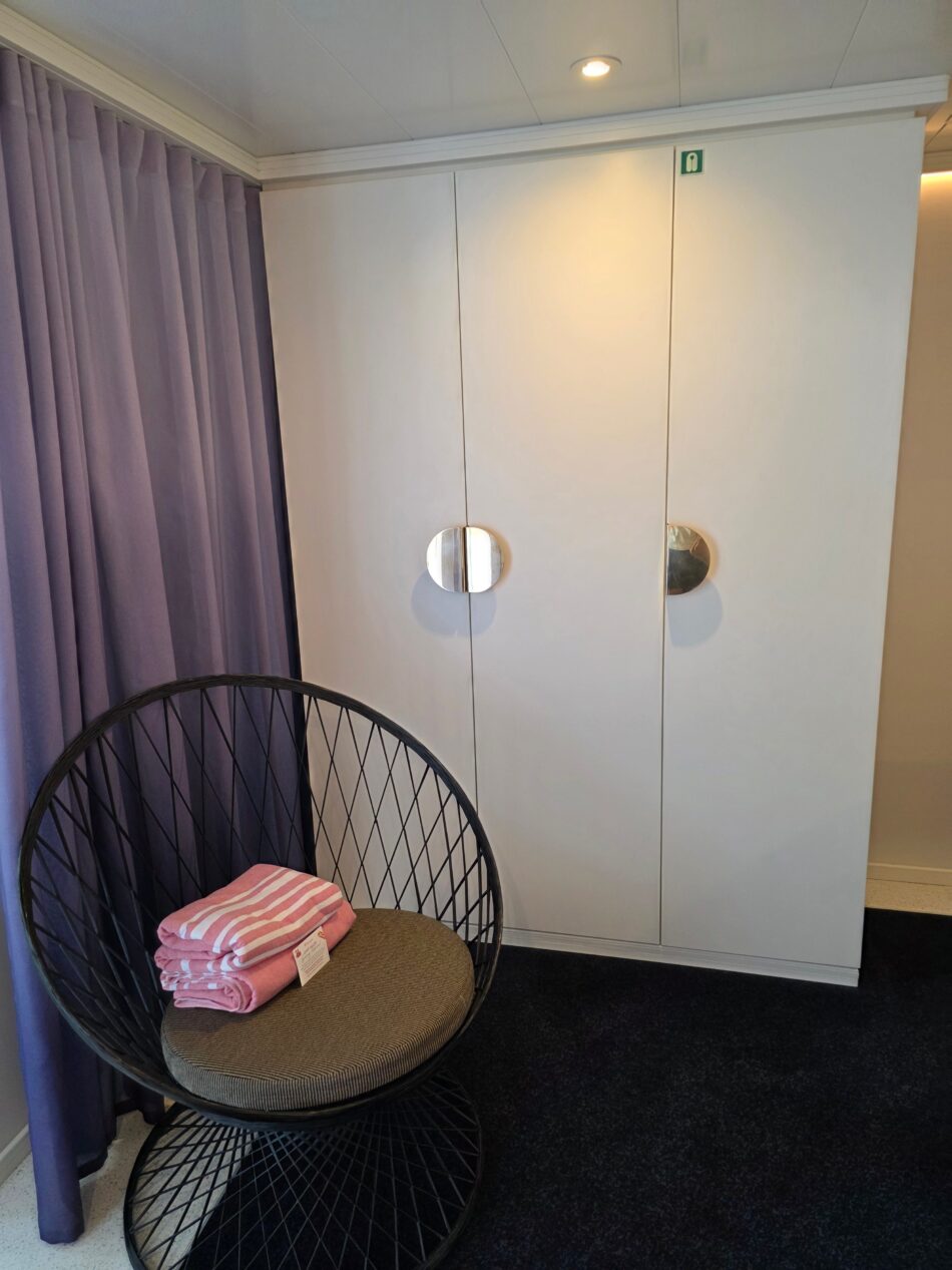 Virgin Voyages cruise ship cabin interior with a modern chair holding folded pink-striped towels, next to a wall with circular handles and purple curtains.